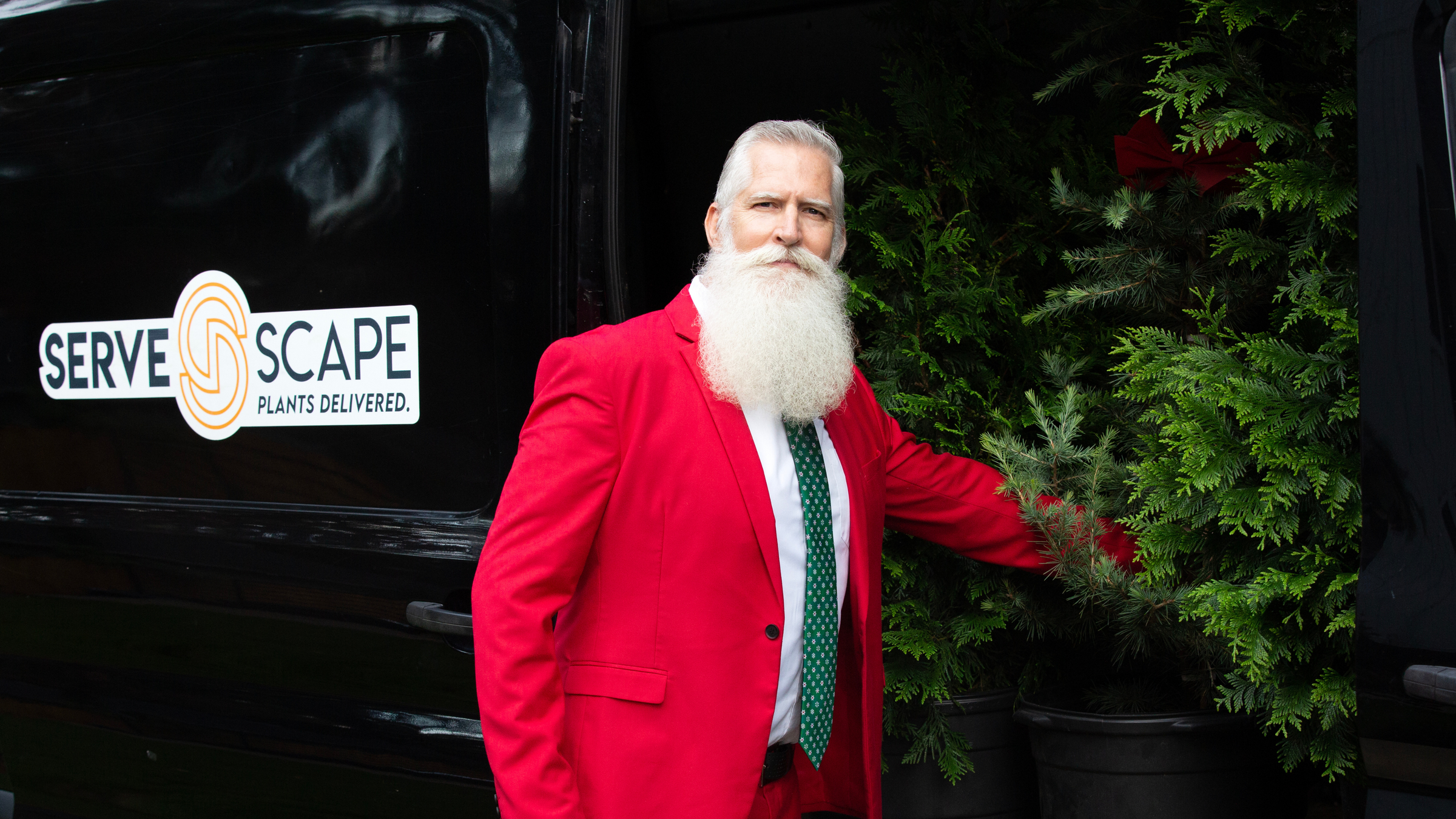 Man in a red suit standing next to a black van with 'ServeScape' branding, holding a potted plant.
